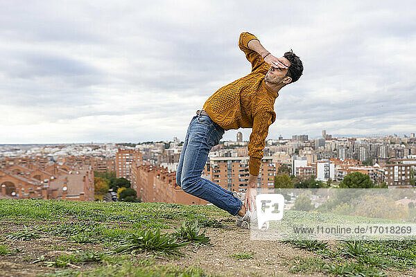 Flexibler junger Mann  der die Augen abschirmt  während er auf einem Hügel in der Stadt gegen den bewölkten Himmel akrobatische Übungen macht