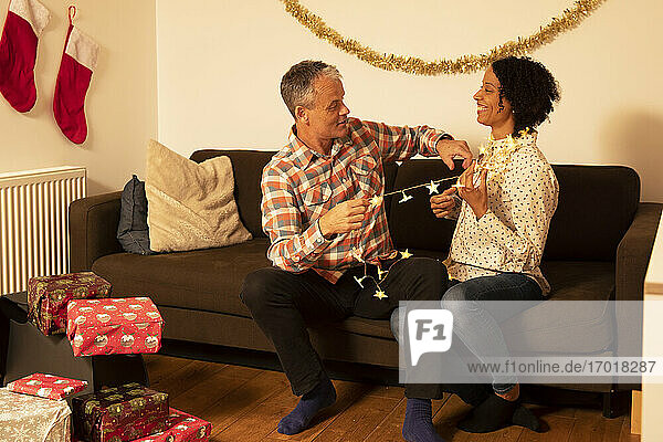 Fröhliches Paar spielt mit beleuchteten Weihnachtslichtern  während es zu Hause auf dem Sofa sitzt