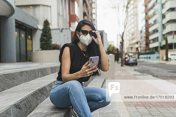 Frau mit Schutzmaske  die auf einer Treppe in der Stadt sitzt und ihr Smartphone benutzt