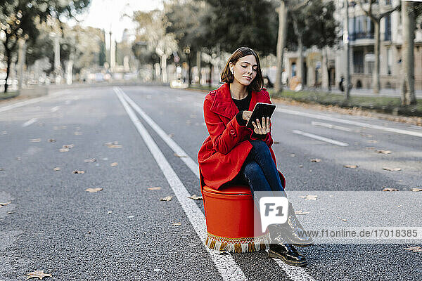Junge Frau in roter Jacke hört Musik  während sie ein digitales Tablet benutzt  das auf einem Sitz auf der Straße steht