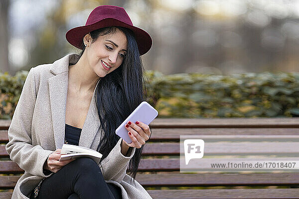 Schöne Frau mit Hut  die ein Mobiltelefon benutzt  während sie auf einer Bank im Park sitzt