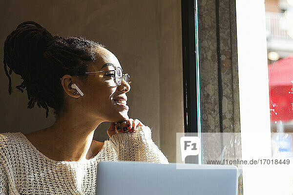 Lächelnde junge Frau  die durch ein Fenster schaut  während sie mit einem Laptop in einem Café sitzt