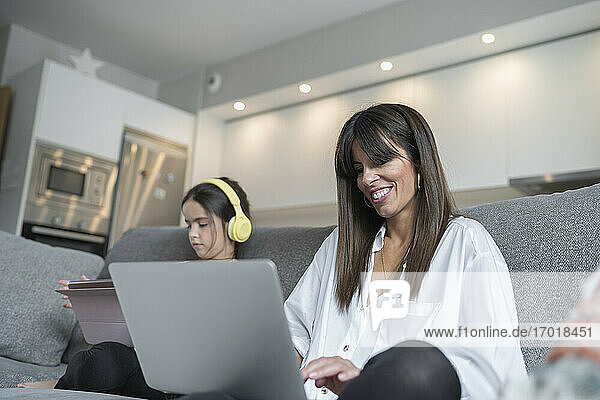 Smiling mother using laptop sitting by daughter holding digital tablet in living room