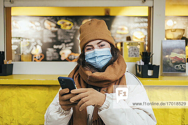 Junge Frau mit Gesichtsmaske und Mobiltelefon im Herbst in einem Café