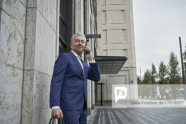 Lächelnder älterer männlicher Unternehmer  der mit seinem Handy telefoniert  während er an einem Gebäude in der Stadt steht