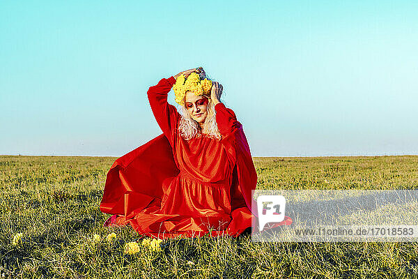 Senior woman putting flowers in hair while sitting on grass against blue sky