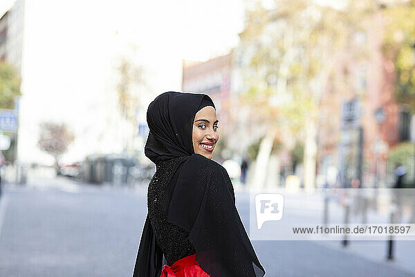 Porträt einer jungen schönen Frau mit schwarzem Hijab  die über die Schulter schaut und in die Kamera lächelt