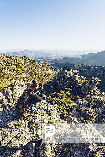 Junge Frau fotografiert männlichen Freund auf einem Berg sitzend gegen den klaren Himmel