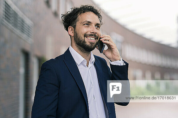 Lächelnder männlicher Unternehmer  der mit seinem Smartphone vor einem Gebäude spricht