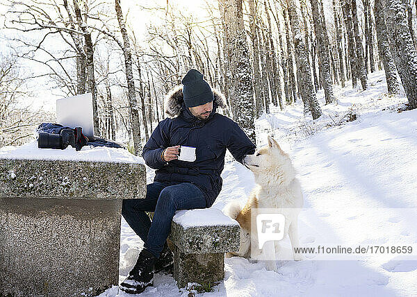 Mid adult man with coffee cup stroking dog while sitting on rock bench in snow