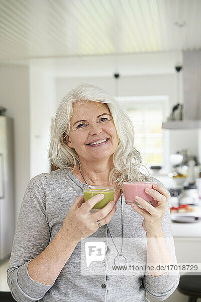 Lächelnde Frau im Ruhestand mit frischen Smoothies in der Hand  während sie in der Küche zu Hause steht