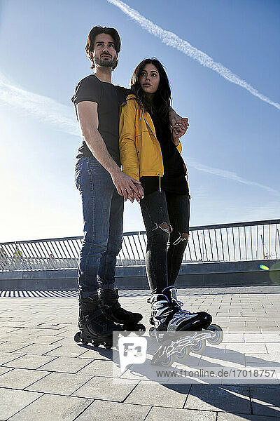 Young man with arm around woman standing on pier against sky during sunny day