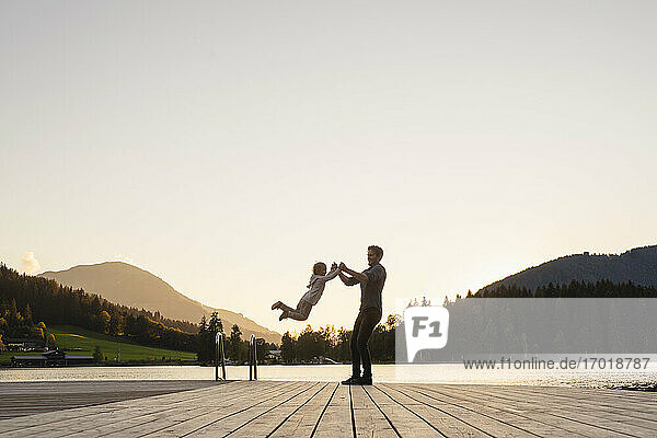 Vater spielt mit seiner kleinen Tochter am Ende des Stegs am Seeufer in der Abenddämmerung