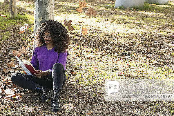 Lächelnde  modische Frau  die ein Buch liest  während sie sich an einen Baumstamm in einem öffentlichen Park lehnt