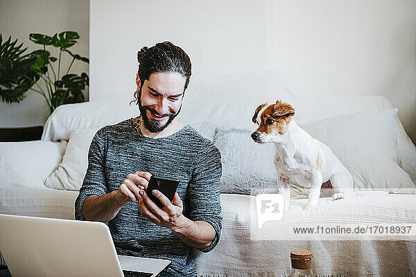 Mann mit Laptop,  der ein Mobiltelefon benutzt,  während er bei seinem Hund zu Hause sitzt