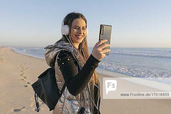 Lächelnde Frau  die ein Selfie mit ihrem Smartphone macht  während sie am Strand gegen den klaren Himmel steht