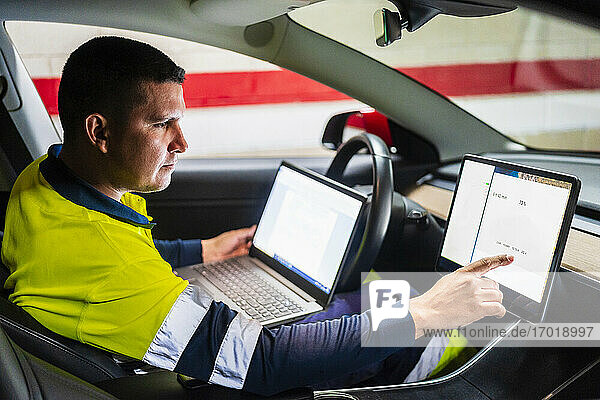 Männlicher Programmierer mit Laptop  der im Elektroauto an einem digitalen Tablet arbeitet