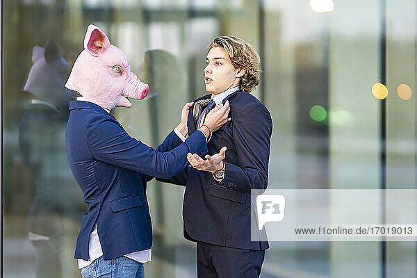 Geschäftsmann mit Schweinemaske hält den Kragen eines männlichen Kollegen an einer Glaswand