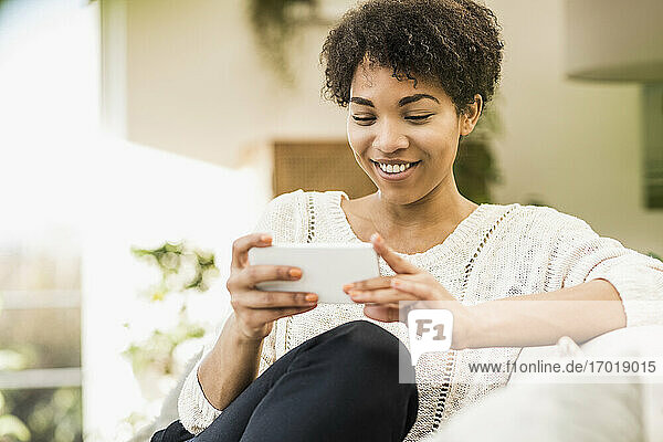Lächelnde Frau  die ein Mobiltelefon benutzt  während sie zu Hause sitzt