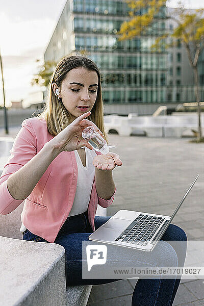 Spain  Barcelona  Young businesswoman with laptop using hand sanitizer outdoors