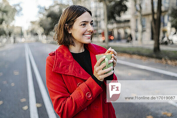Lächelnde junge Frau  die eine Kaffeetasse hält und auf der Straße wegschaut
