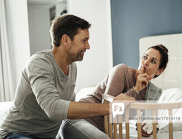 Mature woman with finger on lips looking at man by crib in bedroom