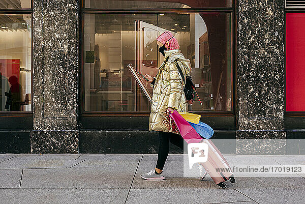 Junge Frau mit Gesichtsmaske  die ein Mobiltelefon benutzt  während sie mit Einkaufstaschen und Gepäck auf dem Gehweg geht