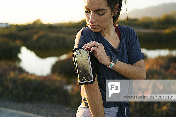 Junge Frau  die ihr Smartphone am Armring einstellt  während sie vor einer Landschaft steht