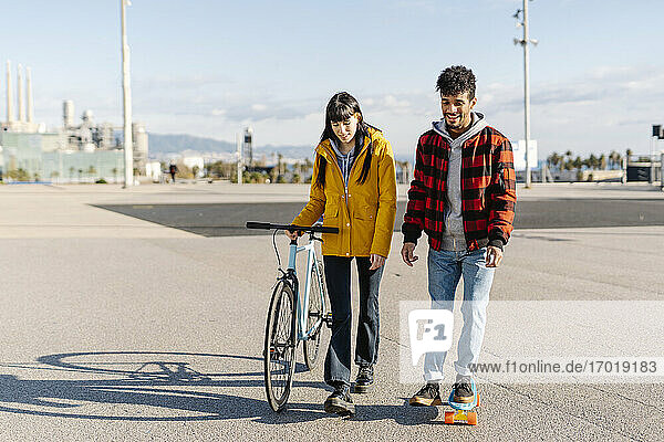 Junge Frau mit Fahrrad geht an einem Freund vorbei  der auf der Straße Skateboard fährt