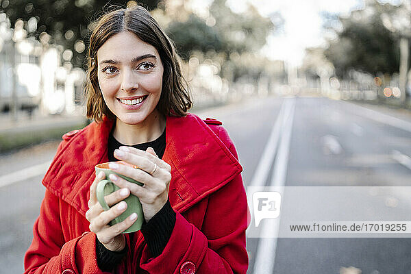Smiling woman in winter jacket day dreaming while holding coffee cup on road