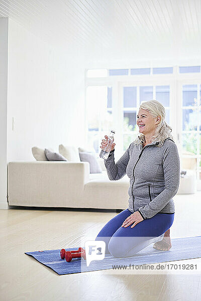 Lächelnde Frau trinkt Wasser  während sie mit Hanteln im Wohnzimmer trainiert