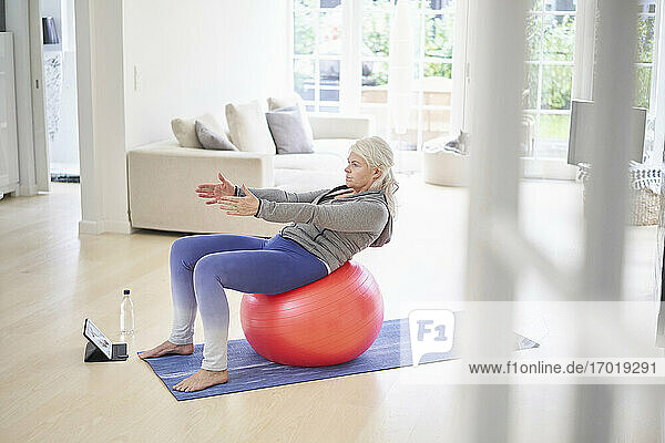 Ältere Frau  die auf einem Fitnessball trainiert  während sie über ein digitales Tablet zu Hause von einem Online-Tutorial lernt