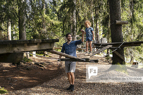 Father assisting little daughter walking across small suspension bridge on forest obstacle course