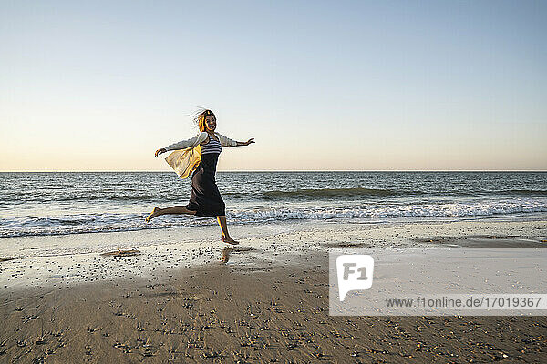 Fröhliche junge Frau läuft am Strand gegen den klaren Himmel bei Sonnenuntergang