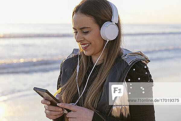 Lächelnde schöne Frau  die ein Mobiltelefon benutzt  während sie bei Sonnenuntergang am Meer steht