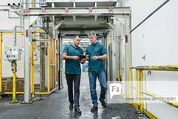 Mature male engineer colleagues walking while discussing over digital tablet in factory