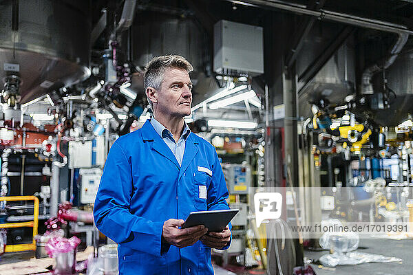 Mature technician day dreaming while holding digital tablet in factory