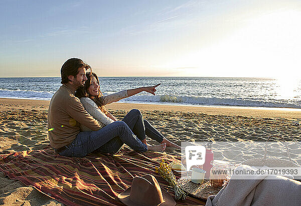 Lächelnde Frau zeigt mit dem Finger auf das Meer  während sie mit einem Mann am Strand sitzt