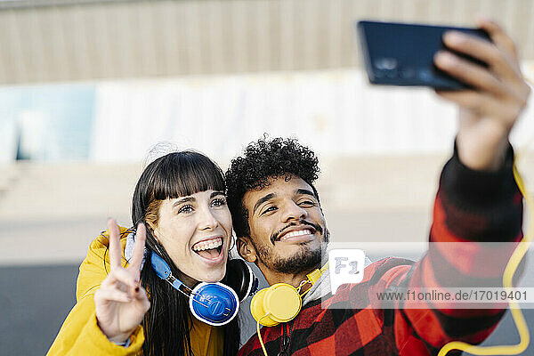 Lächelnder Mann  der ein Selfie über sein Handy mit einem Freund macht  der ein Friedenszeichen zeigt  während er im Freien steht