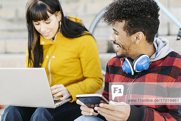 Man with digital tablet smiling while sitting by friend using laptop outdoors