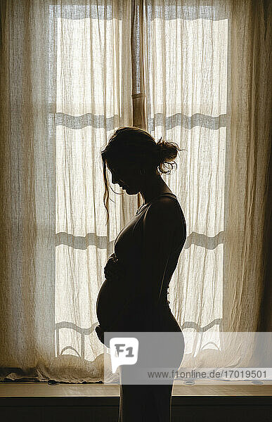Pregnant woman touching belly while standing by window at home