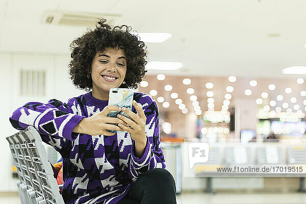 Lächelnde Frau  die ein Smartphone benutzt  während sie am Flughafen sitzt
