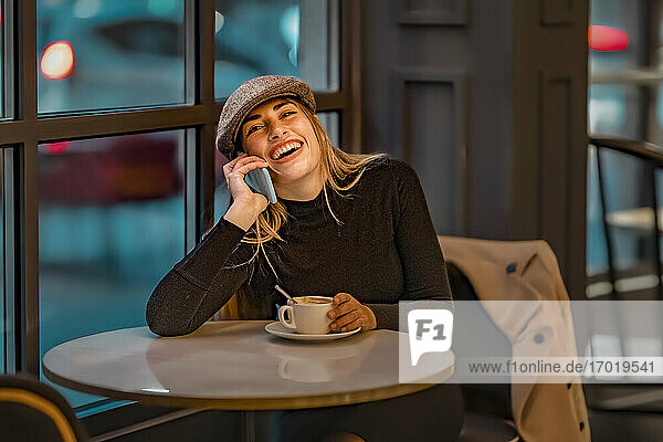 Frau lacht mit Kaffeetasse  während sie in einem Café mit ihrem Smartphone telefoniert