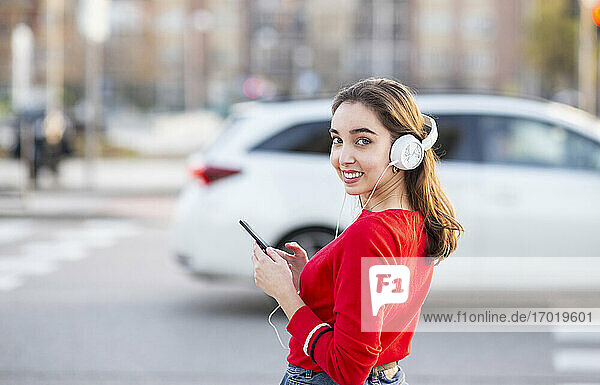 Lächelnde Frau hört Musik über Kopfhörer  während sie auf der Straße in der Stadt steht