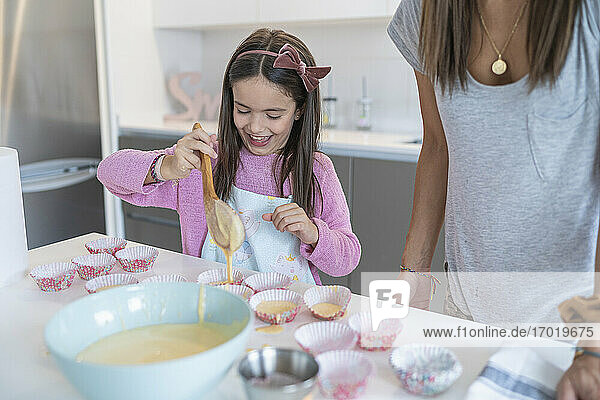 Glückliche Tochter schöpft Teig in Muffinhalter von Mutter in Küche