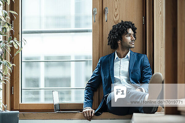 Junger männlicher Berufstätiger schaut weg  während er im Büro am Fenster sitzt und nachdenkt