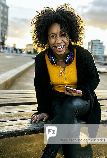 Fröhliche junge Frau mit Afro-Haar sitzt auf einer Bank in der Stadt bei Sonnenuntergang