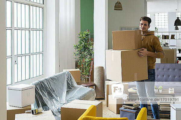 Smiling young man carrying cardboard boxes in living room of new apartment