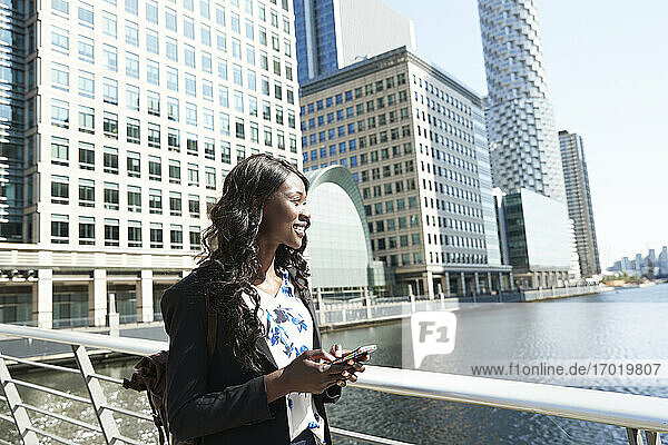 Lächelnde weibliche Fachkraft mit Mobiltelefon  die an einem sonnigen Tag den Fluss betrachtet