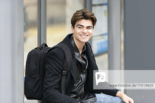 Smiling handsome young man looking away while sitting with backpack at bus stop